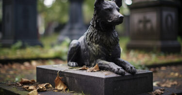 Eine Statue eines Hundes liegt auf einem Sockel umgeben von Herbstblättern auf einem Friedhof.